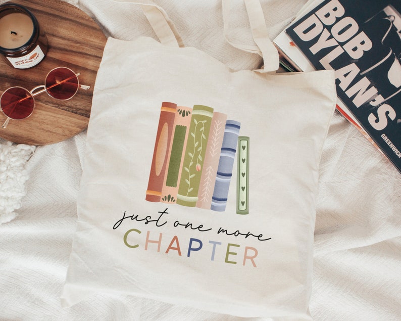 May include: A white canvas tote bag with a design of stacked books with floral details. The text "just one more CHAPTER" is printed in colorful letters below the books.