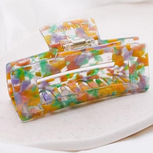 Könnte beinhalten: Ein großer, bunter Haarklammer mit einem Schildpattmuster in Grün-, Gelb-, Rosa- und Lilatönen. Die Klammer hat einen federbelasteten Mechanismus und die Worte "Vida & Shea" sind oben aufgedruckt.