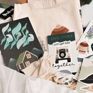 May include: A gift box filled with various items, including a tote bag, a card with a green and blue aurora borealis design, a sticker sheet with a boat and a puffin, and a vinyl sticker with a car and the text "Let's get lost together".