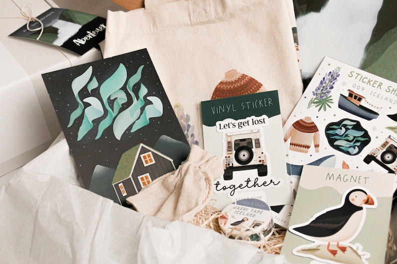 May include: A gift box filled with various items, including a tote bag, a card with a green and blue aurora borealis design, a sticker sheet with a boat and a puffin, and a vinyl sticker with a car and the text "Let's get lost together".
