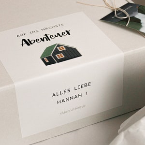 May include: A white gift box with a sticker that says "Auf ins n&auml;chste Abenteuer" and a small illustration of a house. The sticker also says "Alles Liebe Hannah!" and "lilaandthe.de".