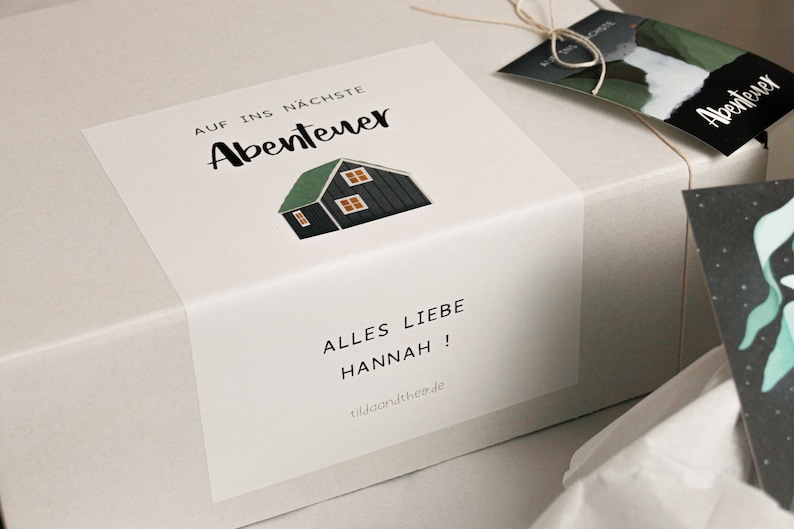 May include: A white gift box with a sticker that says "Auf ins n&auml;chste Abenteuer" and a small illustration of a house. The sticker also says "Alles Liebe Hannah!" and "lilaandthe.de".