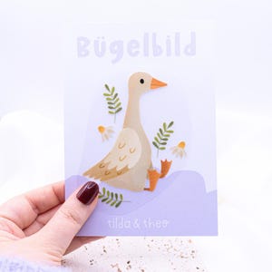Könnte beinhalten: Eine weiße Karte mit dem Text "Bügelbild" in lila Buchstaben. Die Karte zeigt eine Cartoon-Illustration einer Gans mit braunen und weißen Federn, die auf einer lila Welle steht. Die Gans ist von grünen Blättern und weißen Blumen umgeben. Der Text "tilda & theo" ist in schwarz am unteren Rand der Karte gedruckt.