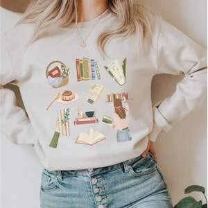 Könnte beinhalten: Ein hellbeiger Pullover mit einem grafischen Druck verschiedener Buch-Motive, darunter Bücherstapel, ein Bücherkorb, eine Tasse Kaffee und eine Frau, die ein Buch liest.