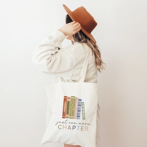 May include: A white canvas tote bag with a stack of colorful books and the text "just one more CHAPTER" printed on it.