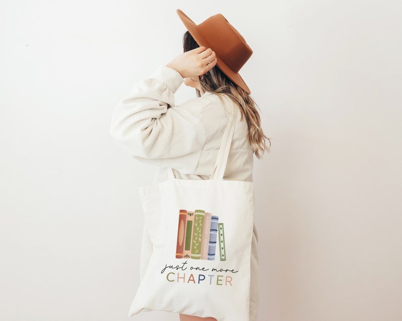 May include: A white canvas tote bag with a stack of colorful books and the text "just one more CHAPTER" printed on it.