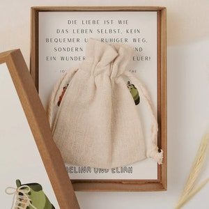 May include: A brown box with a white card inside. The card has the text "Die Liebe ist wie das Leben selbst, kein bequemer ruhiger Weg, sondern ein Wunder" and "Melina und Elian". The box also contains a small white drawstring bag.