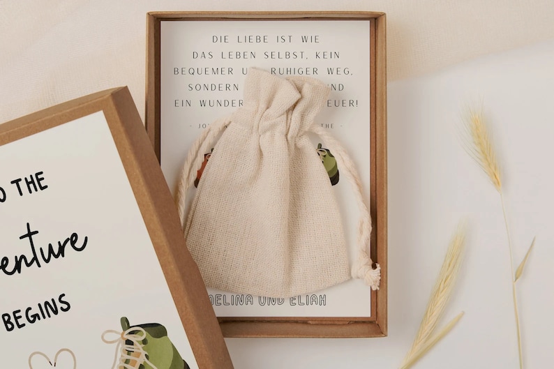 May include: A brown box with a white card inside. The card has the text "Die Liebe ist wie das Leben selbst, kein bequemer ruhiger Weg, sondern ein Wunder" and "Melina und Elian". The box also contains a small white drawstring bag.