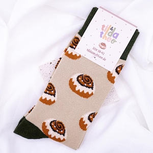 Peut inclure: Une paire de chaussettes beiges avec un talon et une pointe verts. Les chaussettes présentent un motif répété de petits pains à la cannelle bruns avec un glaçage blanc. Les chaussettes ont une étiquette attachée qui dit "tilda & theo" et "Taille 38-42 tildaandiheo.de".