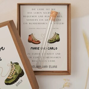 May include: Two brown cardboard boxes with a white background and a quote about love and adventure. The boxes feature illustrations of hiking boots with a heart-shaped string connecting them. The text on the boxes reads "And so the Adventure Begins" and "Die Liebe ist wie das Leben selbst... ein wunderbares Abenteuer!"
