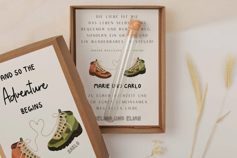 May include: Two brown cardboard boxes with a white background and a quote about love and adventure. The boxes feature illustrations of hiking boots with a heart-shaped string connecting them. The text on the boxes reads "And so the Adventure Begins" and "Die Liebe ist wie das Leben selbst... ein wunderbares Abenteuer!"