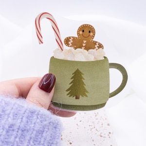 May include: A green mug with a gingerbread man, candy cane, and marshmallows. The mug has a small green Christmas tree on the front.