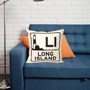 May include: A white throw pillow with a black graphic of a lighthouse and the text "Long Island" on a white background. The pillow is on a blue couch with an orange throw pillow.