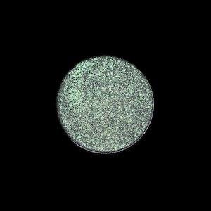 May include: A round, silver-toned eyeshadow pan with a shimmering, glittery, light green surface. The eyeshadow is set against a solid black background, highlighting its texture and color.