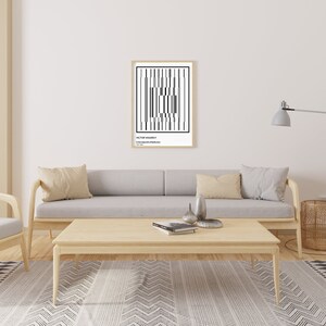 May include: A black and white abstract geometric print in a wooden frame hangs on a white wall above a light gray sofa with beige pillows. A wooden coffee table sits in front of the sofa with a book and two decorative objects on top. A floor lamp with a gray shade and a potted plant are on the right side of the image. A patterned rug is on the floor.