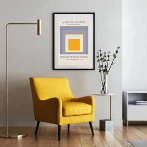 May include: A yellow armchair with black legs sits in a living room with a framed print of a geometric abstract painting by Josef Albers titled "Homage to the Square: Ascending". The print is in a black frame and hangs on a white wall. A gold floor lamp stands to the left of the chair. A small white table with a vase of pink flowers sits to the right of the chair. A white entertainment unit with a black TV sits in the background.
