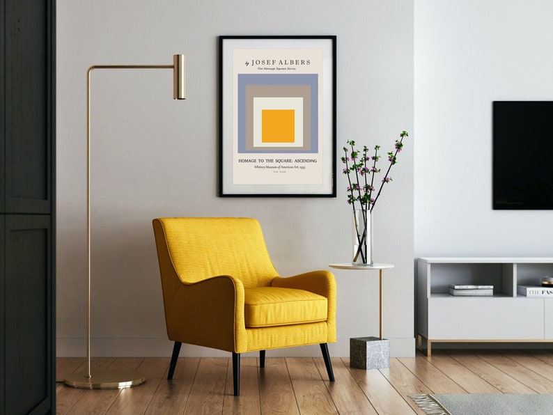 May include: A yellow armchair with black legs sits in a living room with a framed print of a geometric abstract painting by Josef Albers titled "Homage to the Square: Ascending". The print is in a black frame and hangs on a white wall. A gold floor lamp stands to the left of the chair. A small white table with a vase of pink flowers sits to the right of the chair. A white entertainment unit with a black TV sits in the background.