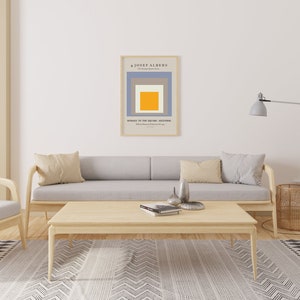 May include: A living room interior with a grey sofa, two beige cushions, a wooden coffee table, a grey armchair, a floor lamp, a potted plant, and a framed print of a geometric abstract art piece by Josef Albers titled "Homage to the Square: Ascending".