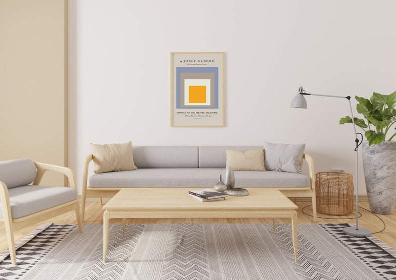 May include: A living room interior with a grey sofa, two beige cushions, a wooden coffee table, a grey armchair, a floor lamp, a potted plant, and a framed print of a geometric abstract art piece by Josef Albers titled "Homage to the Square: Ascending".