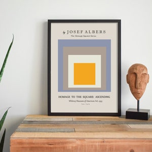 May include: A framed print of a geometric abstract art piece by Josef Albers titled "Homage to the Square: Ascending". The print features a series of squares in shades of blue, grey, and yellow.