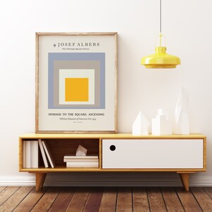 May include: A framed print of a minimalist abstract painting by Josef Albers titled "Homage to the Square: Ascending". The painting features a series of squares in shades of grey, beige, and yellow, with the largest square in the centre being yellow.