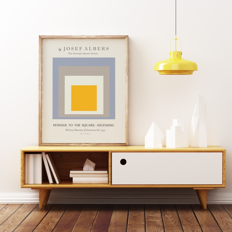 May include: A framed print of a minimalist abstract painting by Josef Albers titled "Homage to the Square: Ascending". The painting features a series of squares in shades of grey, beige, and yellow, with the largest square in the centre being yellow.