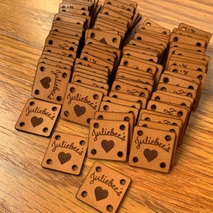 May include: A pile of brown leather labels with the text "Juliebee's" and a heart design. The labels have four holes punched in each corner.