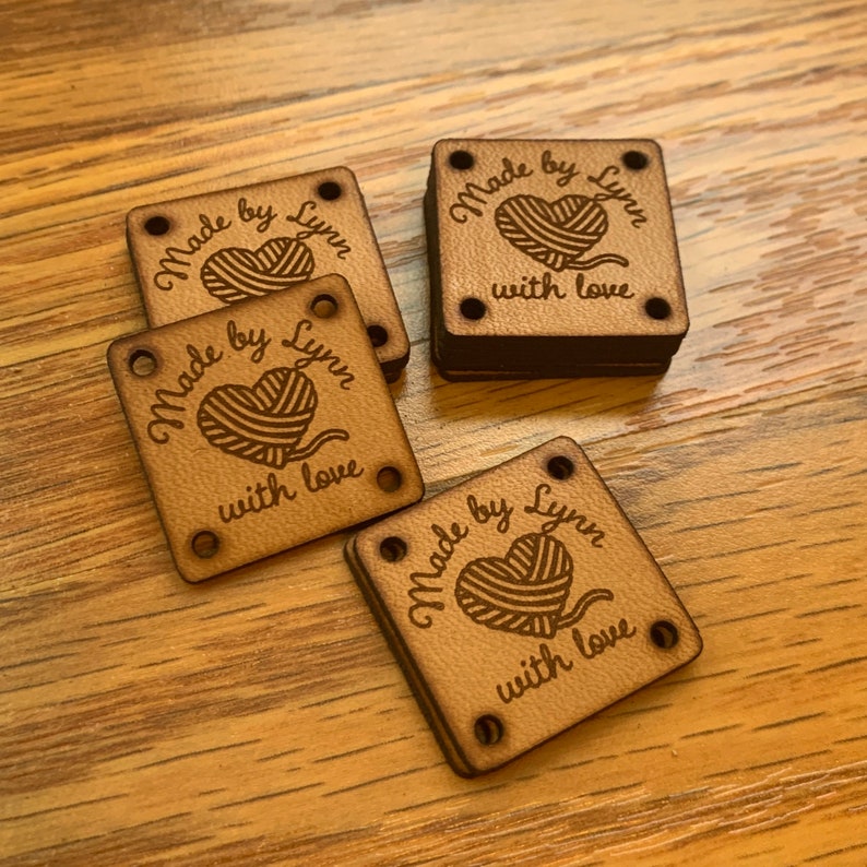 May include: Four brown wooden square tags with rounded corners and four holes. Each tag has the text "Made by Lynn" with a heart-shaped yarn ball graphic and the text "with love".