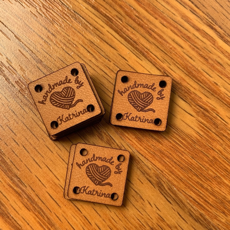 May include: Three brown wooden square tags with rounded corners and holes in each corner. Each tag has the text "handmade by Katrina" with a heart and a ball of yarn inside the heart.
