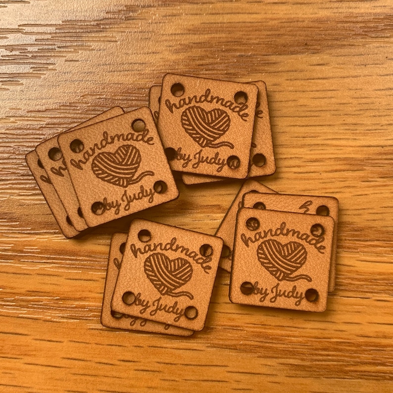 May include: A close-up of six brown leather tags with the text "handmade by Judy" and a heart with a ball of yarn inside. The tags have four holes each.