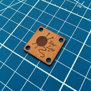 May include: A brown leather square tag with rounded corners and four holes. The tag has a design of a ball of yarn with knitting needles and the text "Your Logo".