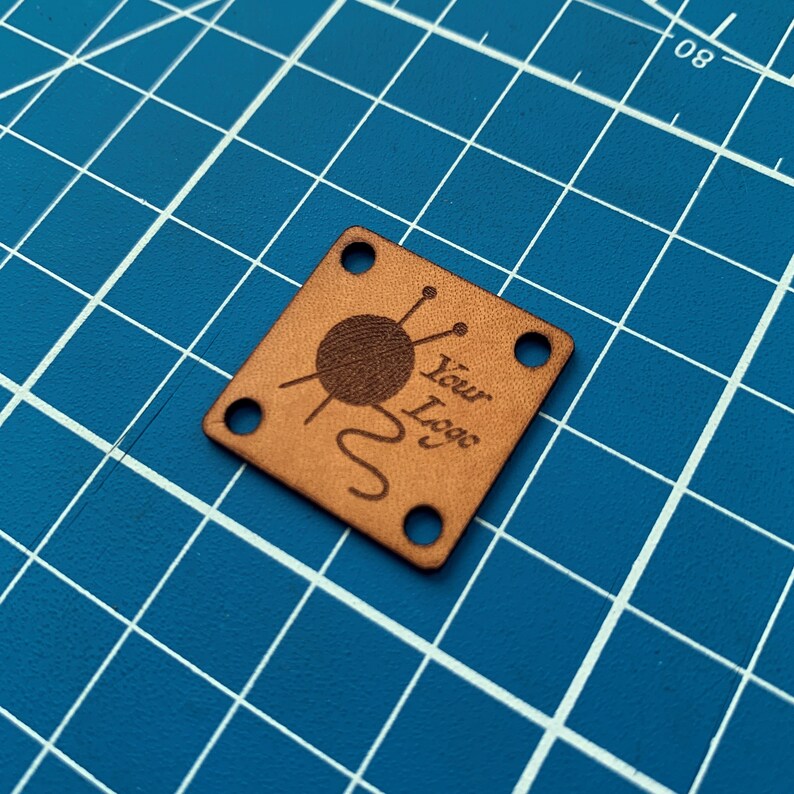 May include: A brown leather square tag with rounded corners and four holes. The tag has a design of a ball of yarn with knitting needles and the text "Your Logo".