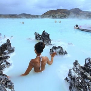 May include: A person wearing a white bikini top relaxes in a milky blue geothermal pool surrounded by black volcanic rocks.