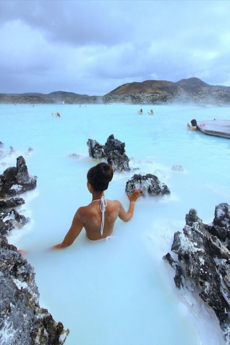 May include: A person wearing a white bikini top relaxes in a milky blue geothermal pool surrounded by black volcanic rocks.