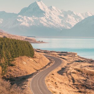 May include: A winding paved road leads towards a snow-capped mountain range in the distance. The road is surrounded by dry, brown grass and a few trees. A body of water is visible in the background.