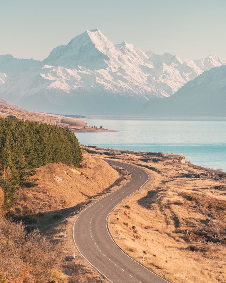 May include: A winding paved road leads towards a snow-capped mountain range in the distance. The road is surrounded by dry, brown grass and a few trees. A body of water is visible in the background.