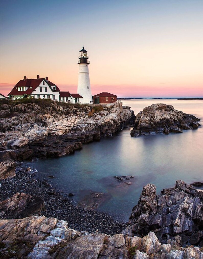 May include: A white lighthouse with a black roof stands on a rocky outcropping overlooking a calm body of water. The sky is a soft pink and blue, creating a serene and picturesque scene.