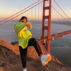 May include: A person wearing a bright green sweatshirt with the word "FLAWED" printed on it, black pants, and white sneakers, standing on a hill with the Golden Gate Bridge in the background.