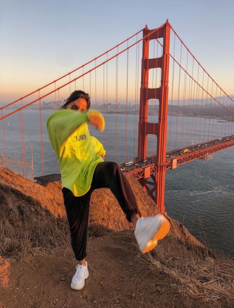 May include: A person wearing a bright green sweatshirt with the word "FLAWED" printed on it, black pants, and white sneakers, standing on a hill with the Golden Gate Bridge in the background.