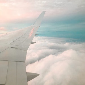 May include: A view from an airplane window, showing the wing of the plane and a vast expanse of fluffy white clouds below. The sky above the clouds is a soft blue with a hint of pink.