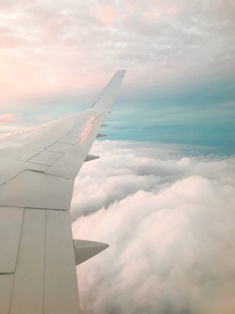 May include: A view from an airplane window, showing the wing of the plane and a vast expanse of fluffy white clouds below. The sky above the clouds is a soft blue with a hint of pink.