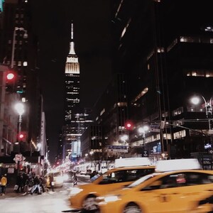 May include: A night view of a city street with yellow taxis driving past a tall skyscraper in the distance.