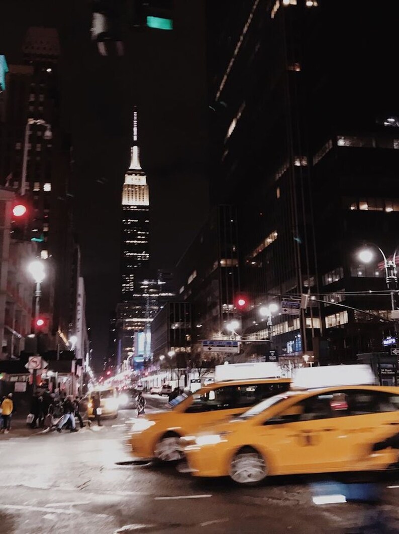May include: A night view of a city street with yellow taxis driving past a tall skyscraper in the distance.