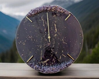 May include: A round, amethyst-colored clock with gold-colored hands and hour markers. The clock face has a dark purple hue with gold flecks, and the outer edge is lined with small, purple crystals. The clock is set on a wooden surface.