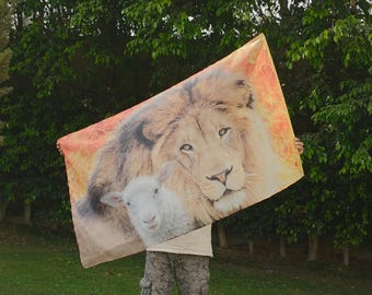 Op de afbeelding: Een rechthoekig wandkleed met een grote, gedetailleerde print van een leeuwengezicht naast een klein wit lammetje. De achtergrond van de print toont warme oranje en gele tinten, wat een vurig effect achter de dieren creëert.