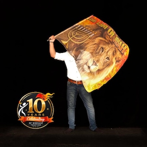 Puede incluir: Una bandera rectangular con un gráfico de un león y una menorá dorada con el texto The Lion Of Judah. La bandera muestra patrones de llamas en tonos naranja y amarillo. Un logotipo circular con el texto 10 Years Celebrating Of Worship Together.