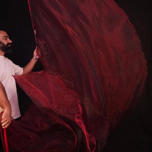 May include: A large, translucent, deep red fabric prop with a shimmering, lightweight texture. The material is captured in motion, creating fluid, sweeping curves and folds that catch the light, displaying varying shades of dark crimson and maroon.