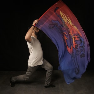 May include: A person in a white t-shirt and grey pants is holding a large, colorful flag with a lion design. The flag is flowing in the air, and the person is looking down at it.
