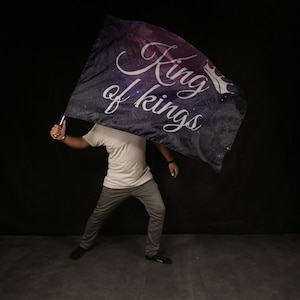 May include: A person holding a black and purple flag with white stars and the text "King of Kings" in a white cursive font.