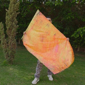 May include: A large rectangular flag featuring a vibrant abstract design with blended shades of orange, yellow, and pink. The fabric has a textured, painted appearance with visible brushstrokes and color gradients throughout the surface.